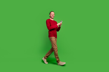 Sticker - Full length photo of good mood smart guy wear red sweatshirt go empty space with smartphone in hands isolated on green color background