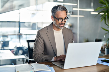 Wall Mural - Smiling mature business man executive wearing suit sitting at desk using laptop. Happy busy professional middle aged businessman ceo investor banker working on computer corporate technology in office.