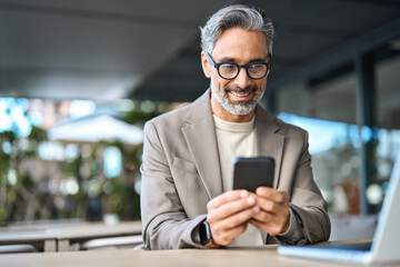 Happy stylish older busy businessman investor holding smartphone looking at cellphone doing banking payments. Middle aged business man using mobile cell phone working outdoors. Candid photo.