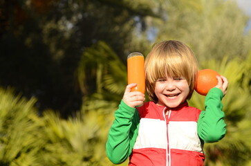 Cheerful child drinks orange juice. A boy in the garden, among the palm trees, holds an orange and a plastic bottle of juice. Healthy juices, vitamins for children, healthy eating