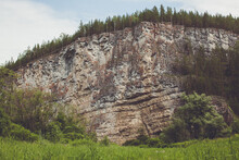 Huge Ancient Rock Cliff Face With Green Trees On The Top And Green Vegetation At The Bottom.