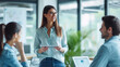 © Studio Nova - A young woman with glasses holding a tablet is standing and engaging in a conversation with seated colleagues in a modern office setting.