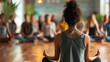 yoga instructor is seen from behind, sitting in a meditative pose with a class of participants in the blurred background
