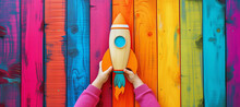 Launch Of A Red Rocket On Colorful Wooden Background, Made Of Wood, Held By Children's Hands. Successful Start Concept.