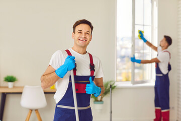 Wall Mural - Portrait of happy smiling young janitor in uniform from professional cleaning service looking cheerful at camera and showing thumb up sign. Housework, household and housekeeping concept.