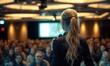 Backview of blonde female long hair with ponytail motivational speaker or coach in front of her conference meeting audience half turned with microphone in her hand