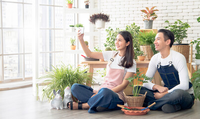 Couple gardener young Asian man woman two person sitting with floor smiling looking hand holding Smartphone selfie camera in calm work shop home plant white wall. hobby job happy and care concept