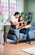 © StockImageFactory - Young and happy Indian couple sitting on sofa at home, using a laptop for shopping or video call.