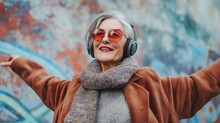 Elderly Woman With Gray Hair Wearing Red Sunglasses And A Brown Coat Smiling And Listening To Music With Headphones Standing In Front Of A Colorful Mural.