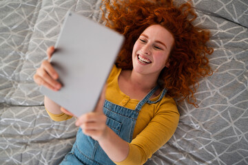 Cheerful confident young woman lying on back indoors and holding a digital tablet. Beautiful girl watching movie alone and laughing.