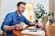 © baranq - Smiling adult man using laptop and phone while freelancing at home