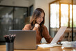 © Wasana - A young professional woman smiles while examining papers in a well-lit, modern office setting, with a laptop.