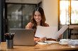 © Wasana - A young professional woman smiles while examining papers in a well-lit, modern office setting, with a laptop.