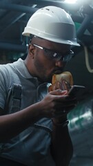 Wall Mural - African American technician eats sandwich and surfs the Internet on mobile phone. Professional engineer in safety uniform and protective hard hat having break working on modern industrial factory.