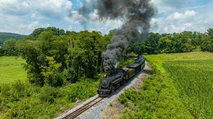 Canvas Print - An Aerial View of a Classic Black Narrow Gauge Steam Train Chugging Along Tracks Surrounded By Lush Greenery And Emitting Thick Smoke Into The Clear Sky.