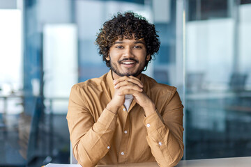 Wall Mural - Smiling Indian businessman in casual attire looking confident at his workplace with a modern office background.