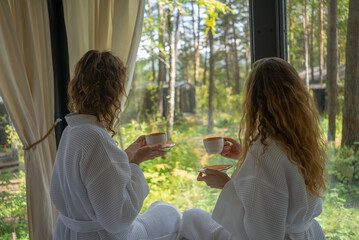 Two women in white bathrobes enjoying coffee together, sitting comfortably with a backdrop of lush green forest visible through a large window, encapsulating a moment of relaxation and companionship