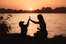 A Black Labrador Retriever Type Mixed Breed Dog Doing A High Five Trick With A Young Woman Silhouetted Against A Sunset At A Lake