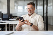 © Stock 4 You - Middle aged Hispanic business manager ceo using cell phone mobile app, laptop. Smiling Latin or Indian mature man businessman holding smartphone sit in office working online on gadget, close up.