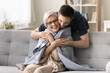 © fizkes - Positive young adult son man caring for happy elderly mature mother sitting on comfortable sofa, throwing warm plaid, scarf on moms shoulders, hugging from behind with love, affection