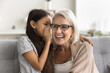 Pretty adorable granddaughter telling secret to happy laughing grandma, whispering at ear. Cheerful grandmother and preteen girl enjoying family communication, listening to kid