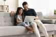 Cheerful young dad and pretty little schoolkid girl using Internet web service on laptop on home couch, watching funny movie, talking on video call, enjoying online communication, entertainment
