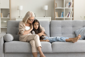 Wall Mural - Serious grandmother and granddaughter kid talking with trust on comfortable couch at home. Caring grandma comforting grandkid, hugging girl with love, warmth, giving compassion