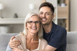 © fizkes - Cheerful caring adult son hugging happy blonde senior mother with love, support, looking at camera with perfect toothy smile, laughing. Elderly mother and grown male child headshot portrait