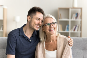 Wall Mural - Happy thoughtful adult child man hugging senior mom with love, care. Dreamy mother and son sitting on sofa at home, looking away, smiling, enjoying meeting, leisure, discussing family plans