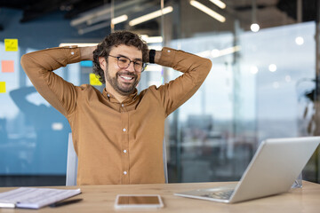 Well done businessman successfully completed work project inside office at workplace, man with hands behind head resting happily smiling , looking at laptop screen satisfied with achievement results