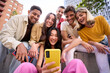 © CarlosBarquero - Young group of gen z people having fun using cell phone together outside watching something funny at mobile app. Cheerful community of student enjoying social media content. Multiethnic friends