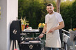 © Aleksandr - Portrait of handsome young chief in apron stand by the BBQ grill and smile. Male look into camera and rubs hands.