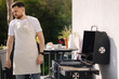 © Aleksandr - Side view of handsome man try how hot is BBQ grill. Man in apron make dinner outdoor