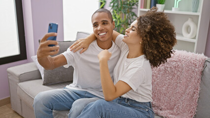 Wall Mural - Beautiful couple enjoying fun lifestyle, sitting on sofa, making confident expressions, taking selfie picture with smartphone at home. smiling, joy-filled love together in their living room.