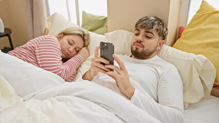 Sticker - A man using a smartphone while a woman sleeps beside him in a cozy bedroom setting, depicting a modern couple's lifestyle.