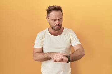 Wall Mural - Middle age man with beard standing over yellow background checking the time on wrist watch, relaxed and confident