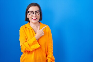 Wall Mural - Middle age hispanic woman wearing glasses standing over blue background cheerful with a smile of face pointing with hand and finger up to the side with happy and natural expression on face