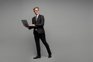 Full body side view adult employee IT business man corporate lawyer wears classic formal black suit shirt tie work in office hold use laptop pc computer look aside isolated on plain grey background.