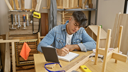 Canvas Print - Hardworking young hispanic man masterfully navigating carpentry business, deftly taking notes on touchpad amid timber and furniture in busy workshop