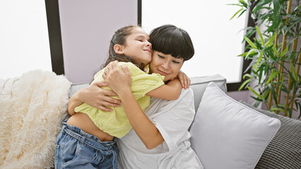 Wall Mural - Confident mother and daughter sharing a joyful hug, smiling heartily while sitting on the living room sofa at home â€“ a beautiful expression of family love and happiness