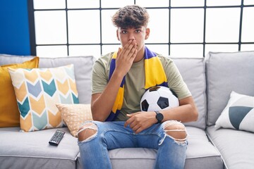 Wall Mural - Hispanic teenager sitting on the sofa watching football match covering mouth with hand, shocked and afraid for mistake. surprised expression