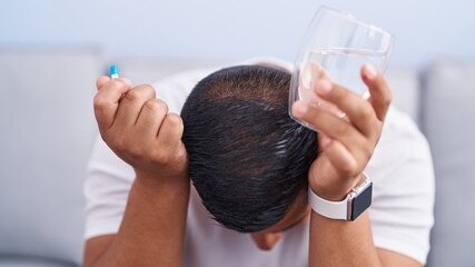 Poster - Young latin man stressed sitting on sofa holding pill and glass of water at home