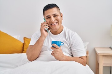 Wall Mural - Young latin man talking on smartphone drinking coffee at bedroom