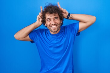 Sticker - Hispanic young man standing over blue background posing funny and crazy with fingers on head as bunny ears, smiling cheerful