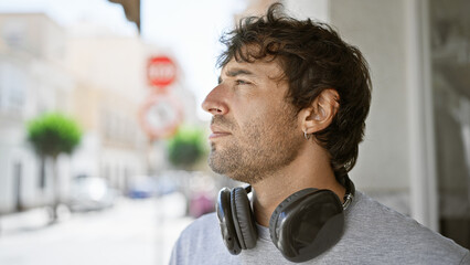 Poster - Cool, serious young guy with blond beard, rocking an urban lifestyle, looking to the side with a concentrated expression, wearing headphones outdoors in sunny street