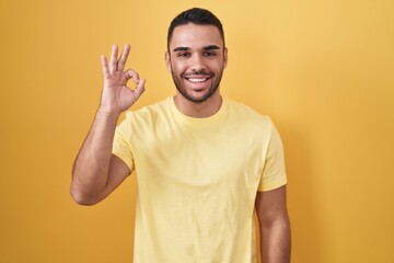 Poster - Young hispanic man standing over yellow background smiling positive doing ok sign with hand and fingers. successful expression.