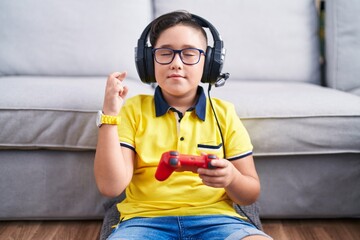 Canvas Print - Young hispanic kid playing video game holding controller wearing headphones gesturing finger crossed smiling with hope and eyes closed. luck and superstitious concept.