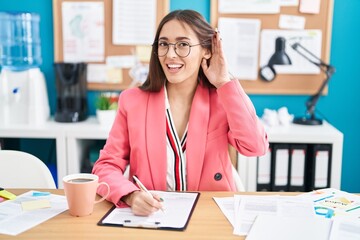 Canvas Print - Young hispanic woman working at the office wearing glasses smiling with hand over ear listening an hearing to rumor or gossip. deafness concept.