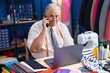 Middle age grey-haired man tailor talking on smartphone using laptop at tailor shop