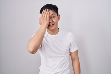 Sticker - Young asian man standing over white background covering one eye with hand, confident smile on face and surprise emotion.